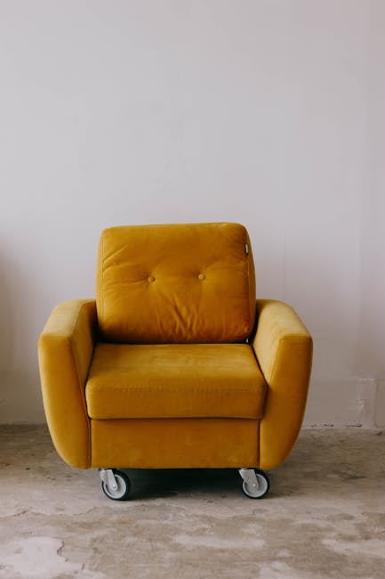 A mustard-yellow upholstered armchair with a modern, rounded design, positioned on a concrete floor against a plain white wall. The chair features a tufted backrest with two visible button details and